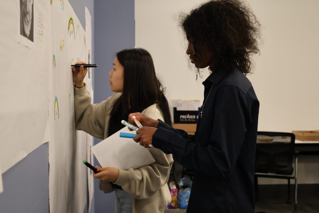 A student writing on a giant piece of paper on the wall next to another student